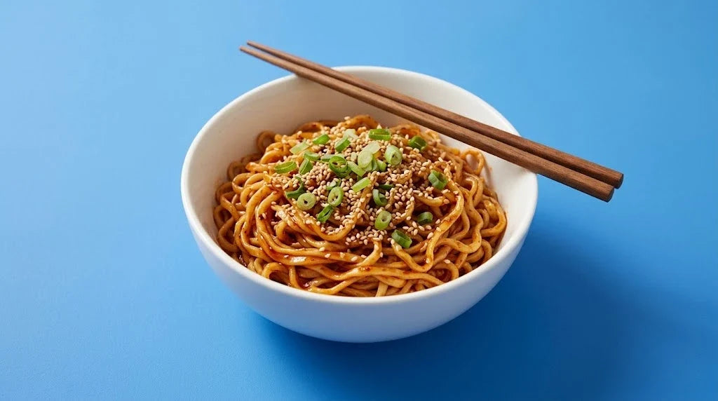A bowl of noodles in red chili oil with scallions and sesame, chopsticks on top, on a blue background