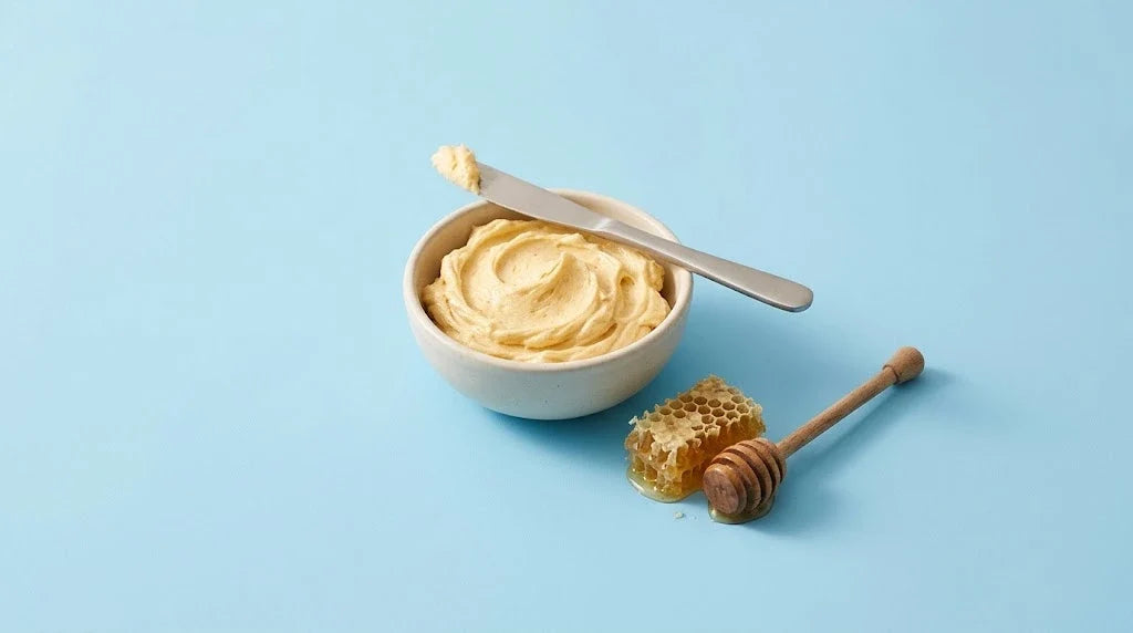 A bowl of whipped honey butter with a knife, honeycomb and a honey dipper on a sky-blue background