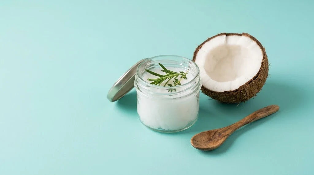 A jar of white coconut oil with rosemary and thyme, half a coconut and a wooden spoon on a turquoise background
