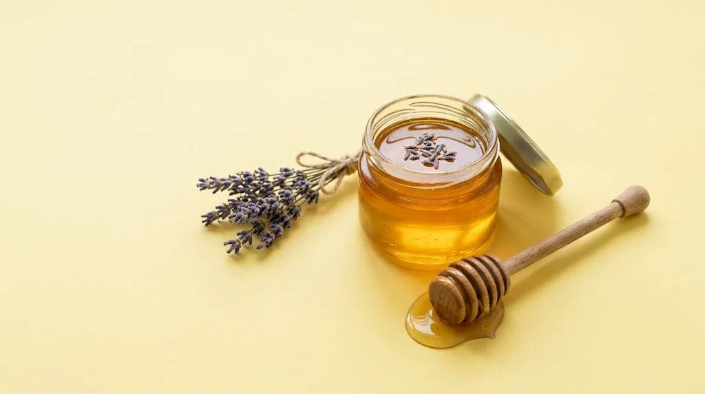 A jar of honey topped with lavender buds, a honey dipper and dried lavender on a butter-yellow background
