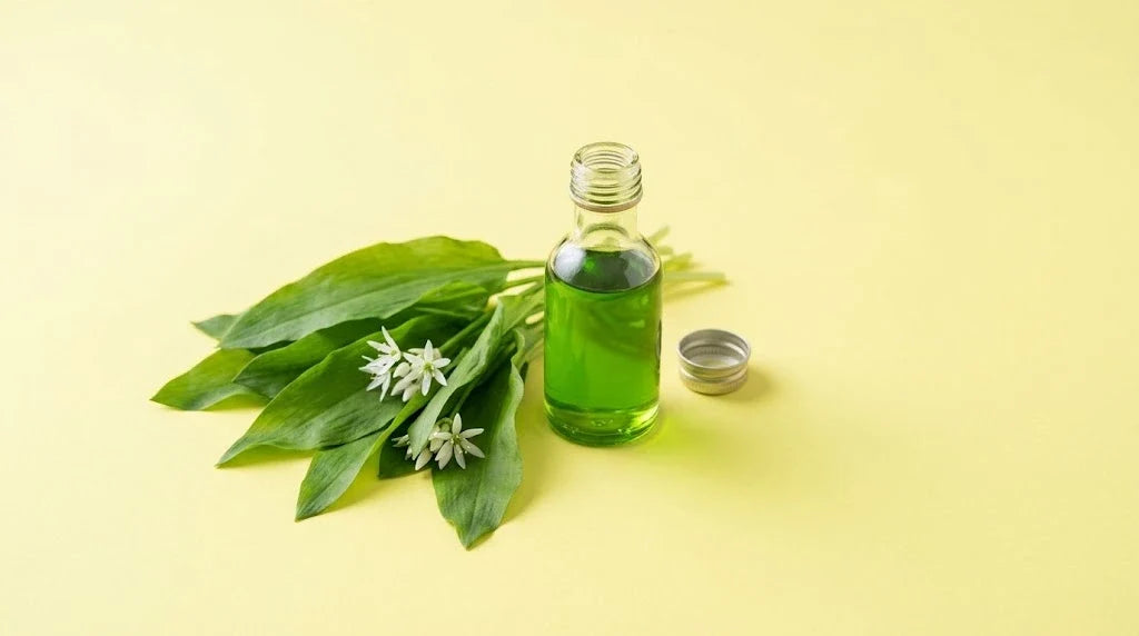 A bottle of bright green wild garlic oil beside fresh wild garlic leaves and flowers on a pale yellow background